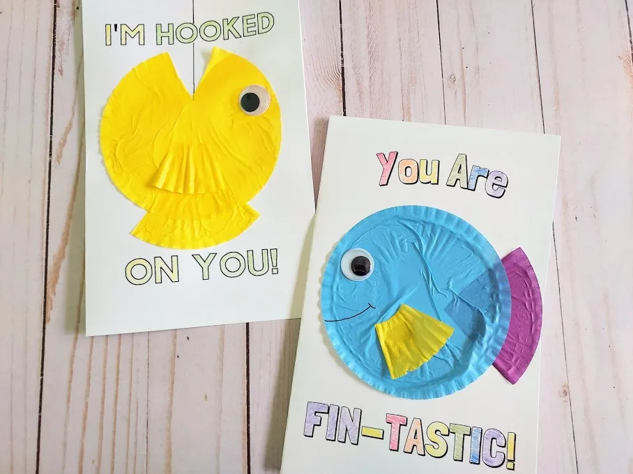Two finished fish card crafts laying next to each other. Left card says I'm Hooked On You with a yellow cupcake liner fish with open mouth cut out and aimed at top of card. A black marker line drawn from top of card into the open fish mouth. Right card says You Are Fin-Tastic with a multicolored cupcake liner fish in the center.