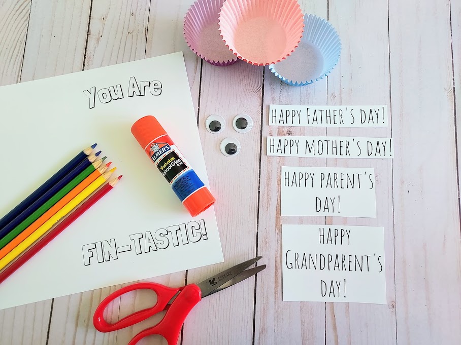 Supplies for fish card craft laying on table includes printable card, color pencils, scissors, glue stick, googly eyes, cupcake liners, and inside holiday phrases.