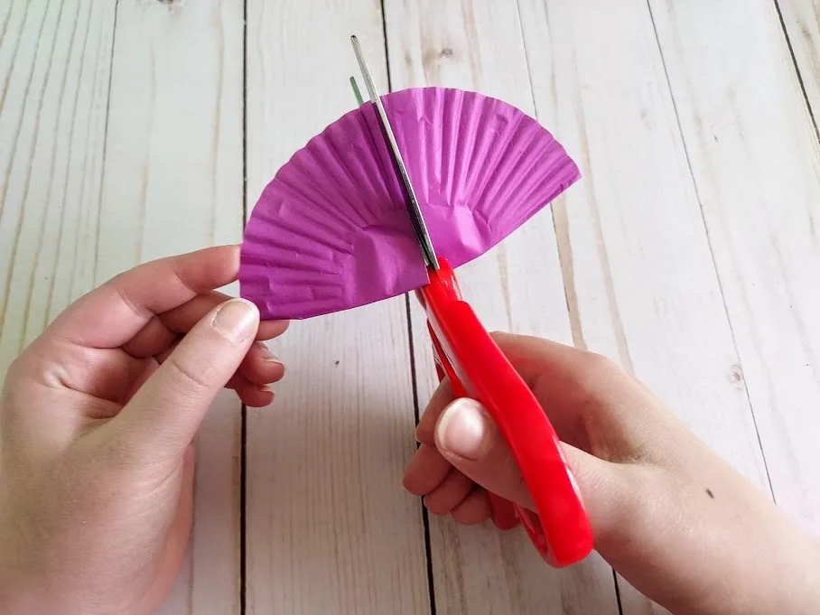 Close view of white child's hands holding and cutting folded purple cupcake liner with scissors.