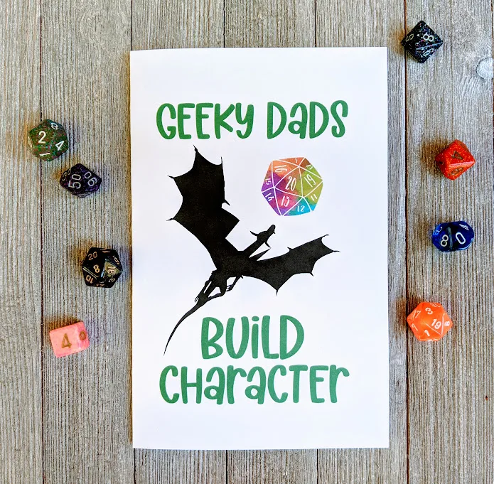 Overhead view of card printed out. Front of card has Geeky Dads in green text at the top, then a dragon silhouette flying towards a rainbow watercolor D20, and below the dragon is green text Build Character. Assorted polyhedral dice are scattered around the card. 