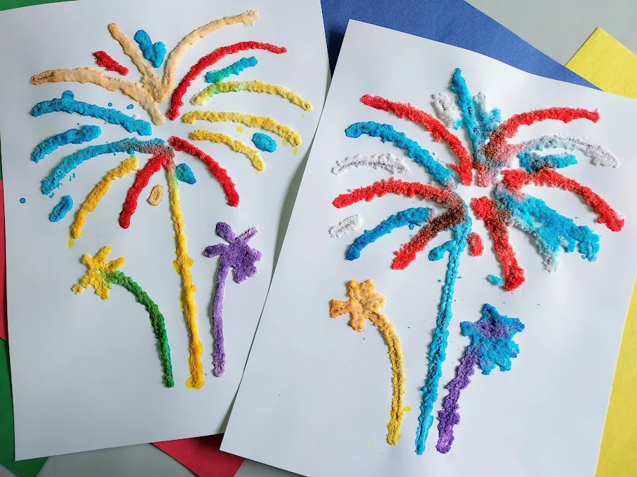 Two completed fireworks salt paintings using red, blue, yellow, purple, and orange watercolors. They lay on top of assorted colors of construction paper.