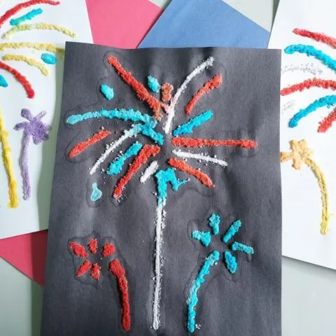Three completed salt paintings of fireworks laying next to each other, slightly overlapped, on top of red and blue construction paper. Middle one is done on black construction paper using blue and red paints. The other two are on white paper and use a colorful variety of paints.