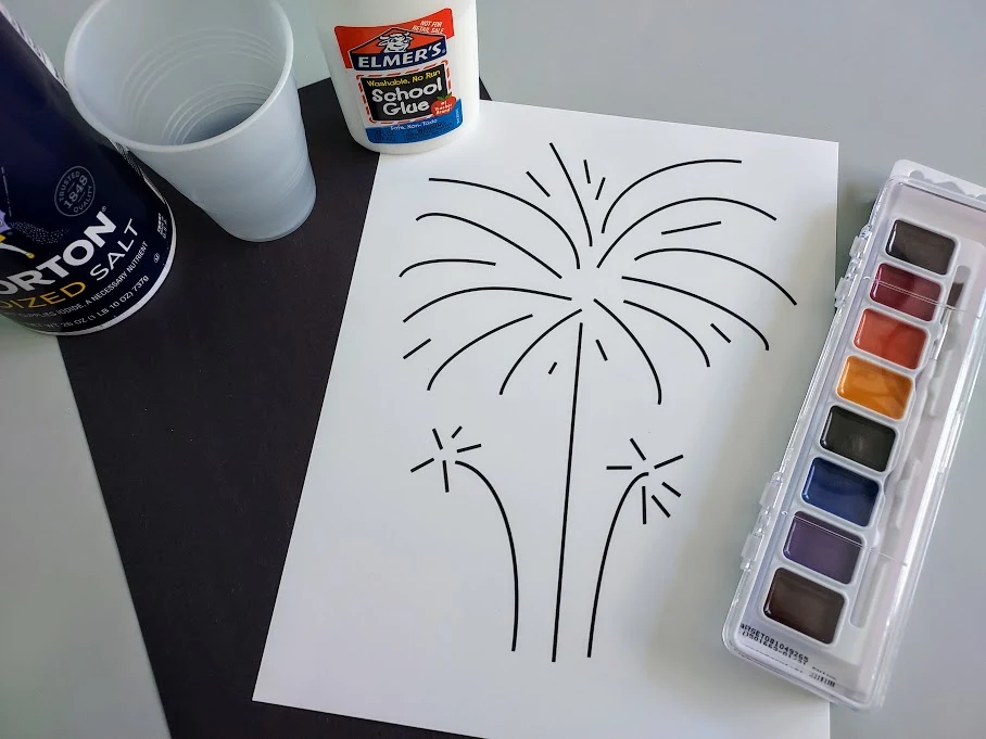Overhead view of printed out fireworks template on white cardstock, a piece of black construction paper, water colors, liquid glue, cup of water, and container of table salt.