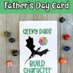Black and white text on a green background at top says Printable Father's Day Card. Photo of printed out card that says Geeky Dads Build Character with a dragon and watercolor D20. Assorted dice are placed around the card.