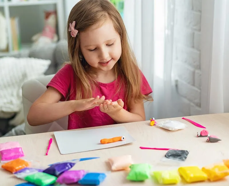 White girls with long brown hair sitting at a desk rolling modeling clay or playdough between her hands. Assorted colors of modeling clay on the desk.