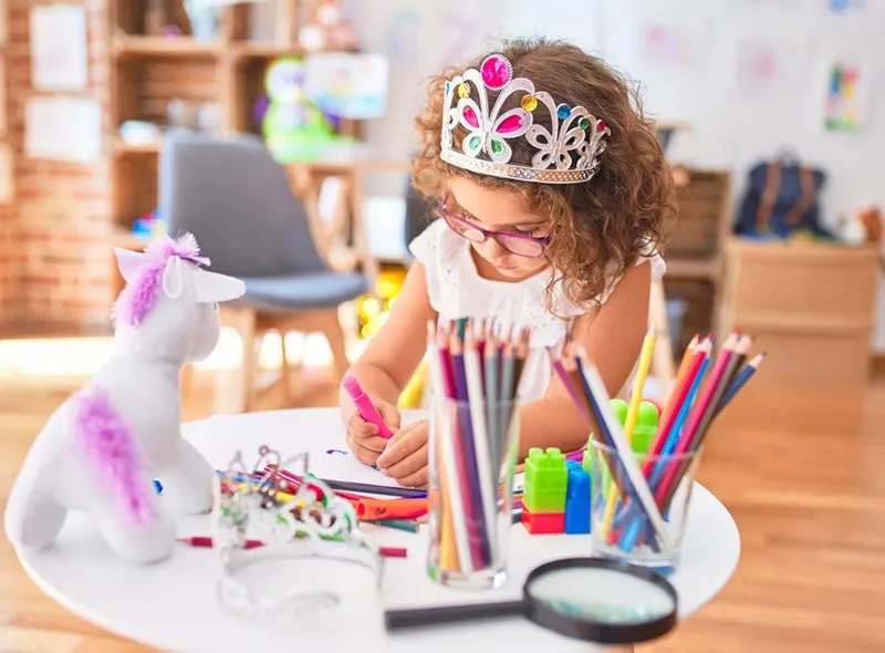 Young girl with curly brown hair, wearing glasses and a princess crown drawing at a table full of markers, colored pencils, magnifying glass, and a few toys.