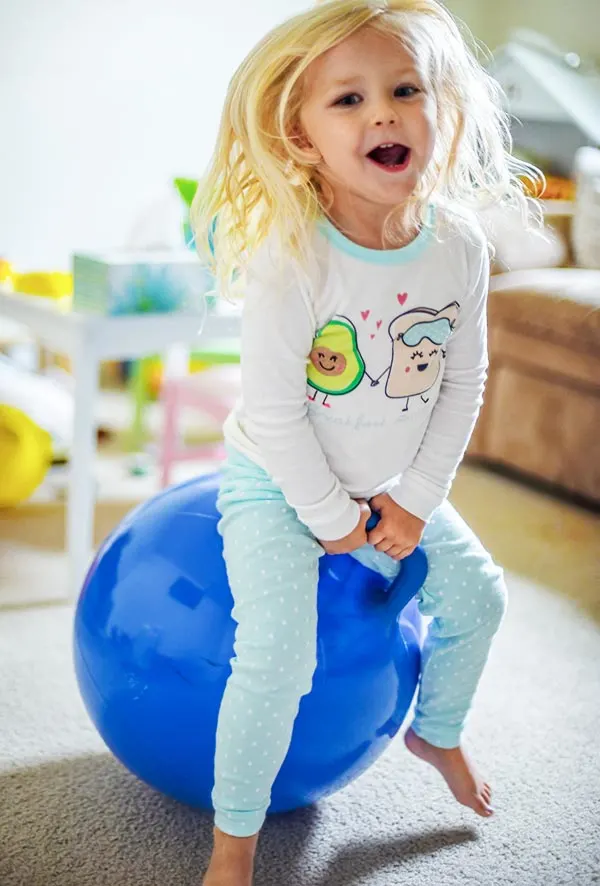 Young white blonde girl smiling and bouncing on a blue hopper ball toy in playroom.
