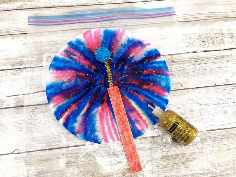 Blue and red colored coffee filter with red colored craft stick glued to it. Gold chenille stem sticking out the top of the craft stick with blue pom pom on the end. Coffee filter laying on top of ziptop bag. Gold glitter glue used to make swirl designs.