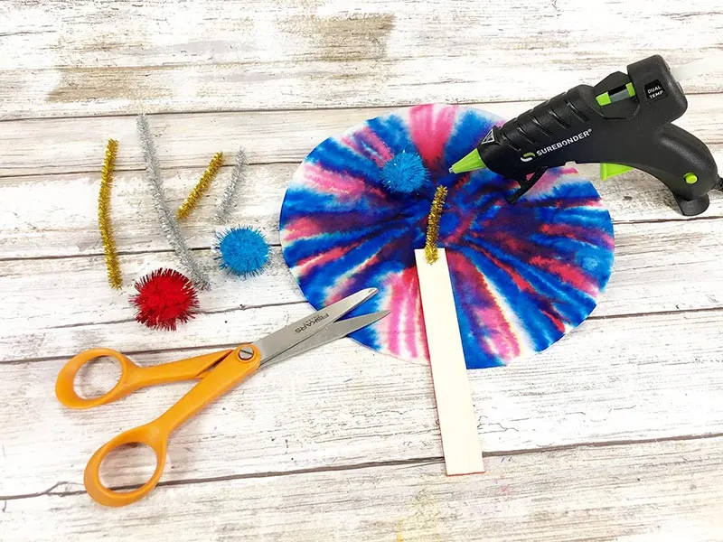 Hot gluing chenille stem to back of colored craft stick and gluing pom pom to end of chenille stem.