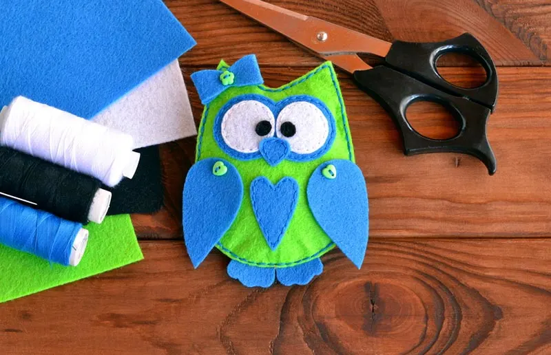 Cute hand sewn green and blue felt owl in the middle. Pieces of felt, thread, and fabric scissors lay around the owl.