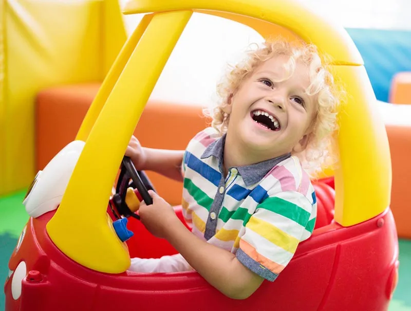 White preschool child with curly blonde hair laughing while inside red and yellow coupe car riding toy.