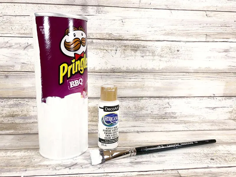Empty Pringles can partially painted over with white paint standing upright next to a bottle of white acrylic paint and a paint brush.