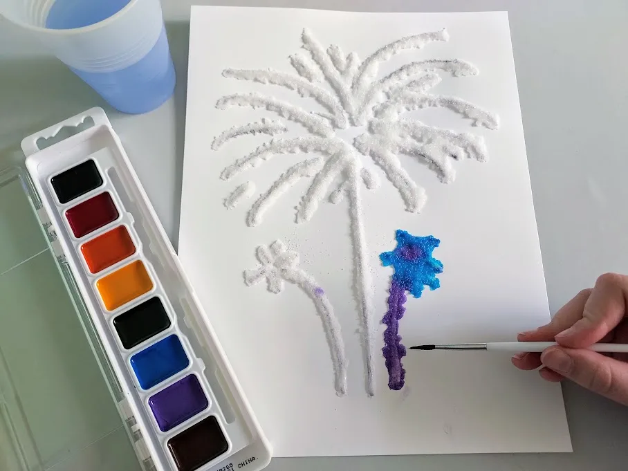 Overhead view of white girl's hand holding paintbrush and dabbing watercolor paints onto salt that's been glued to white cardstock paper in fireworks design.