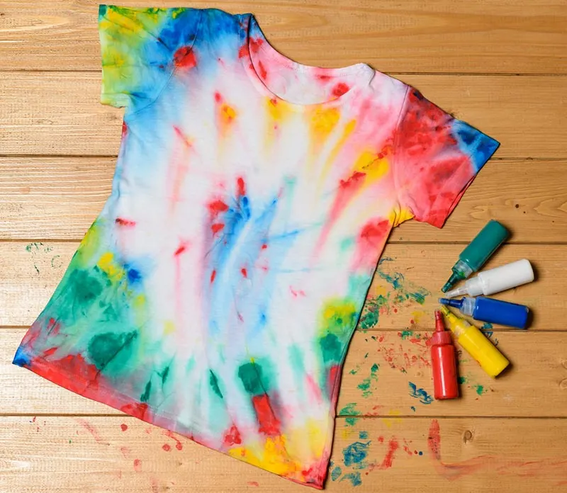 Tshirt laying flat on wooden floor painted in tie dye style. Squeeze bottles of paint laying next to shirt and paint splatter on floor around shirt.
