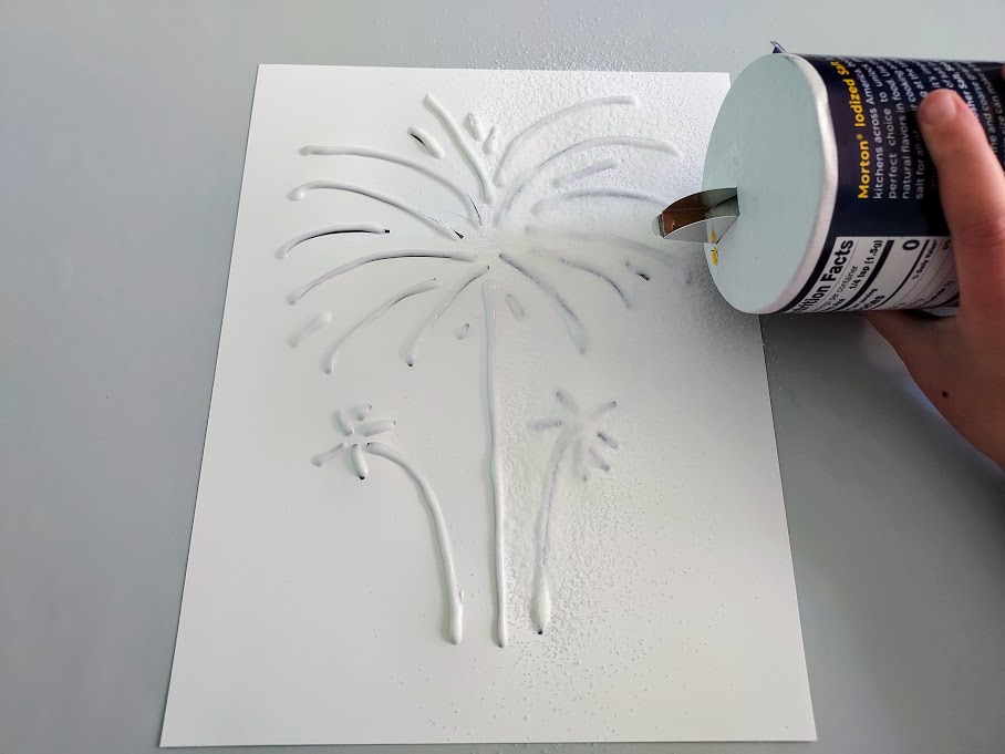 Kid pouring table salt over paper with fireworks glue lines on it.