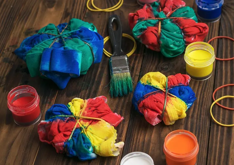 Shirts and clothing items wrapped in rubber bands and colors applied to create tie dye style items. Open paints and a paint brush on the table between clothing pieces.