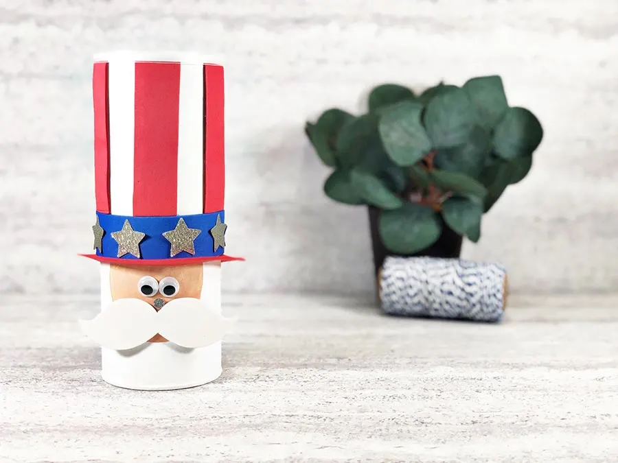Finished Uncle Sam chip can Fourth of July craft for kids. Chip can made to look like Uncle Sam's hat and face in a cute style. Off to the right in the back is a small fake green plant and a roll of twine.