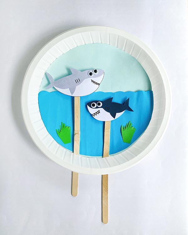 Overhead view of completed swimming sharks paper plate craft project. Gray shark is above wavy water line and navy shark is swimming below the surface.