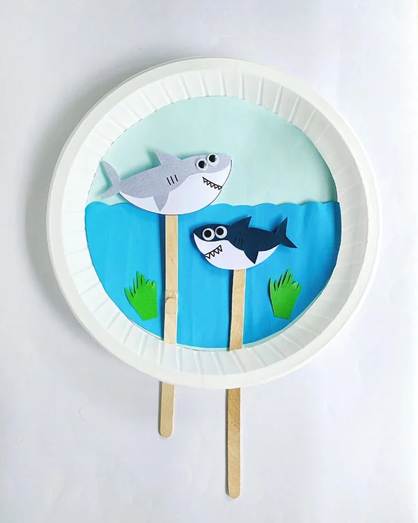 Overhead view of completed swimming sharks paper plate craft project. Gray shark is above wavy water line and navy shark is swimming below the surface.