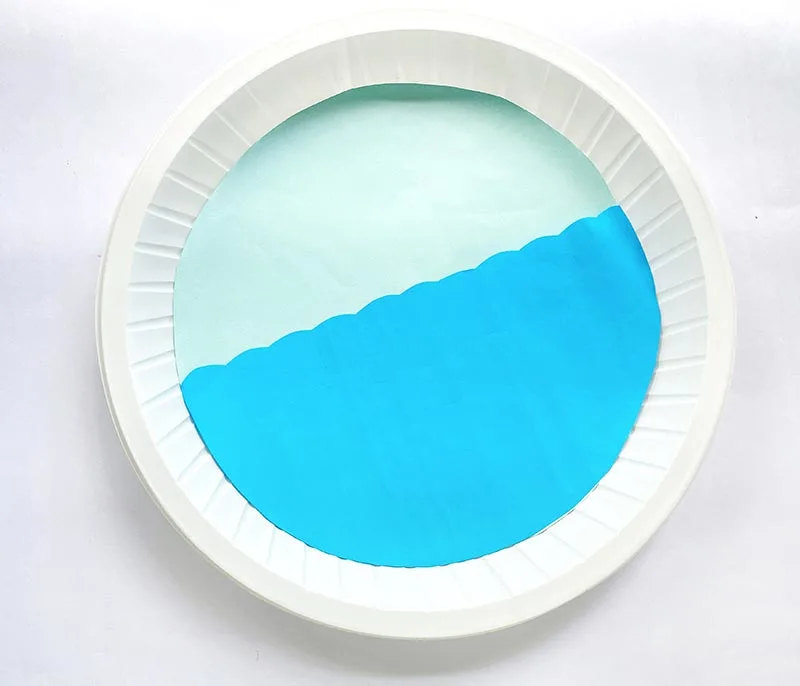 Light blue paper circle glued to center of plate with darker shade of blue cut in semi-circle with wavy line across glued on top of light blue paper to create the ocean.