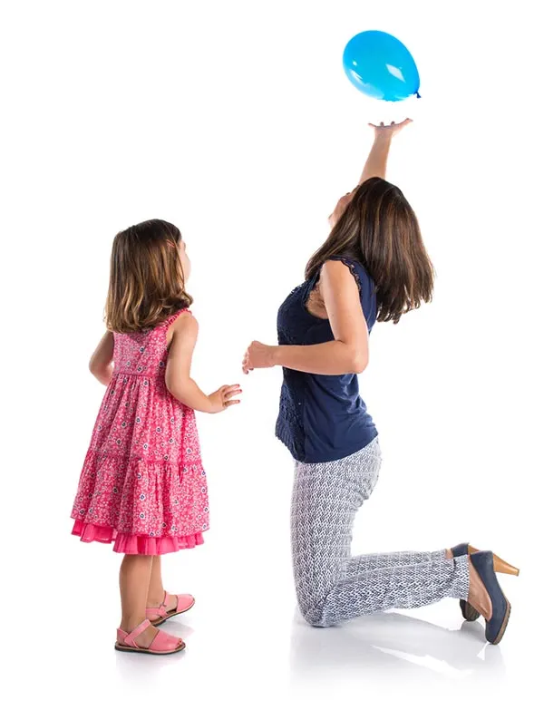 Young white girl with dark hair wearing a pink dress standing and watching as her mom (white woman with short dark hair wearing a navy tank top, light patterned pants, and high heels) kneels in front of her trying to keep a blue balloon in the air.