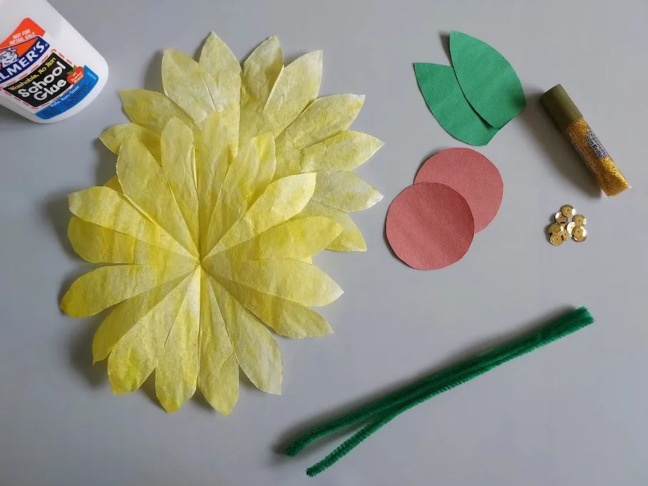 Yellow dyed coffee filters with petals cut, brown paper circles, green paper leaves, sequins, glitter glue, green pipe cleaners, and glue.