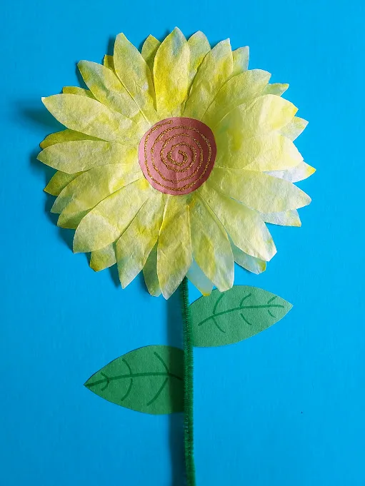 One finished coffee filter sunflower on blue paper with green stem and leaves. Center of flower is a brown construction paper circle with a glitter glue swirl.