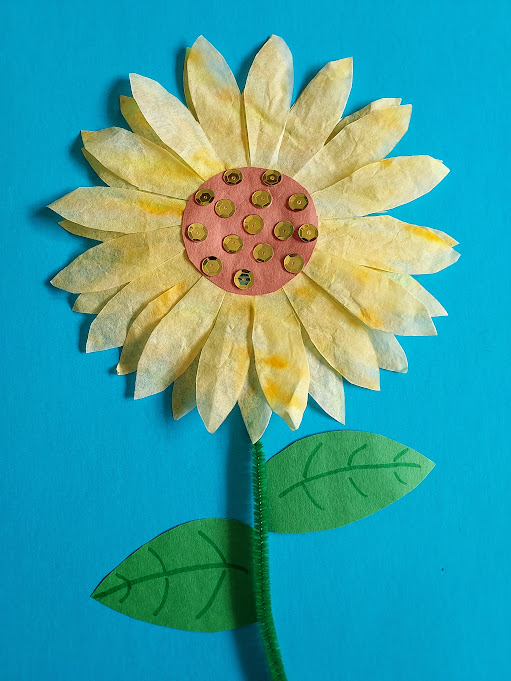 Completed sunflower made with coffee filters, construction paper, chenille stem and sequins glued to blue construction paper.