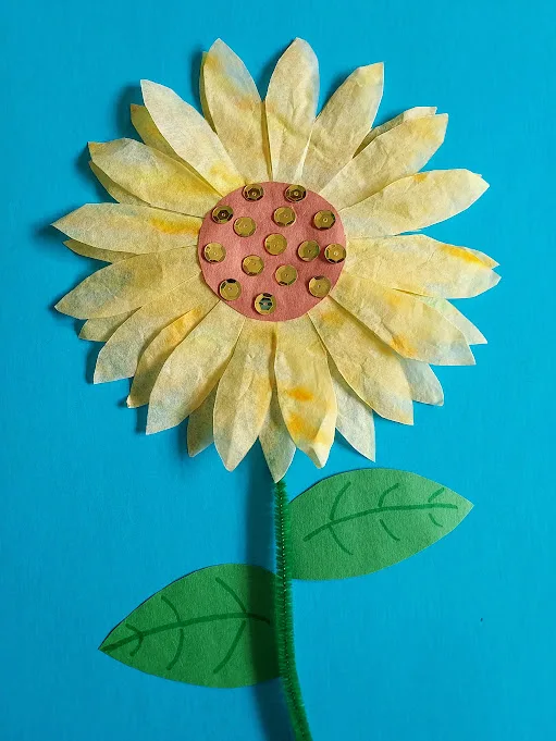 Completed sunflower made with coffee filters, construction paper, chenille stem and sequins glued to blue construction paper.