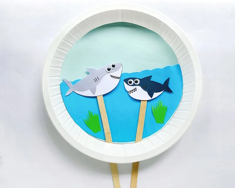 Overhead view of moving shark paper plate craft. Two paper shark puppets (one gray and white, the other navy and white) on popsicle sticks through a slit in a paper plate. Blue paper glued to back of plate to look like water.