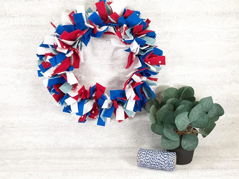 Fourth of July wreath made with red, white, and blue felt strips hanging on a white brick wall next to a small green decorative plant and a roll of twine.