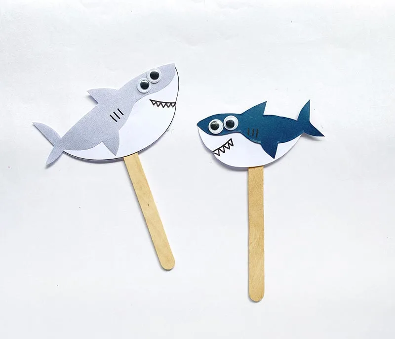Two paper sharks (one gray with white and the other dark navy with white) attached to craft popsicle sticks to make shark puppets.