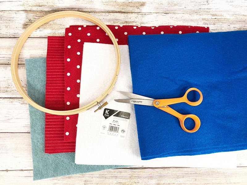 Overhead view of supplies for July felt wreath craft. Squares of light blue, red, red with white polka dots, white, and dark blue felt, wooden embroidery hoop, and scissors laying on top of the felt.