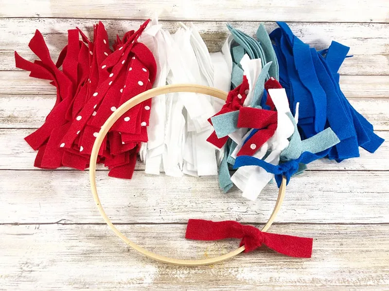 Overhead view of wooden hoop with a few felt strips tied onto it laying partially on top of piles of felt strips sorted by red, white, and blue colors.