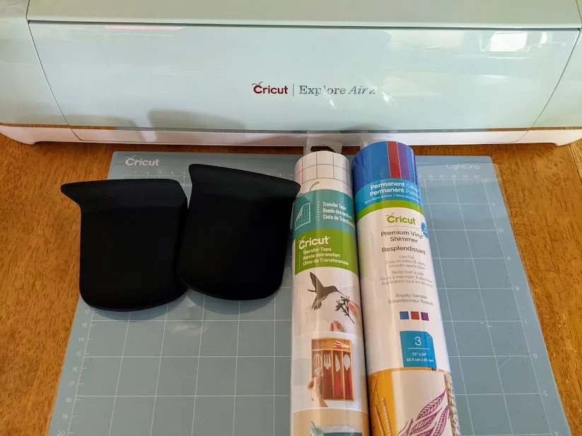 Cricut Explore Air 2 cutting machine, LightGrip machine mat, Premium Shimmer Vinyl, roll of Transfer Tape, and two black headphone hangers on the table for this project.