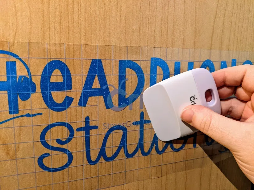 White woman's right hand using white and lavender scraper to apply blue shimmer vinyl design to the side of a wooden cabinet.