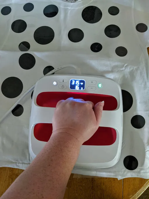 White woman's left hand using EasyPress 2 to apply black iron on vinyl spots to white shirt.