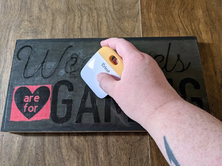 White woman's hand using scraper tool to apply vinyl to black and red painted wood sign.