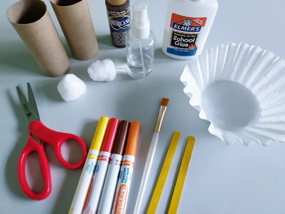 Empty toilet paper tubes, cotton balls, scissors, markers, craft sticks, paint brush, brown paint, spray bottle, glue, and coffee filters spread out on top of off white craft mat.
