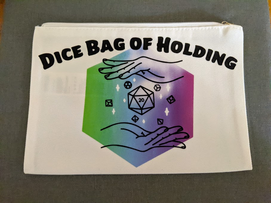 Overhead view of zippered cosmetics bag with custom design applied to it using an EasyPress. Black text at top of bag says Dice bag of Holding above black outline of a pair of hands with floating dice between them. Design outline is overlaid on an infusible ink rainbow hexagon.