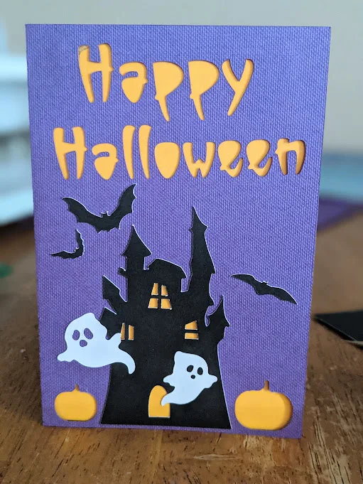 Completed haunted house Halloween card standing up on table in front of Cricut Maker 3.