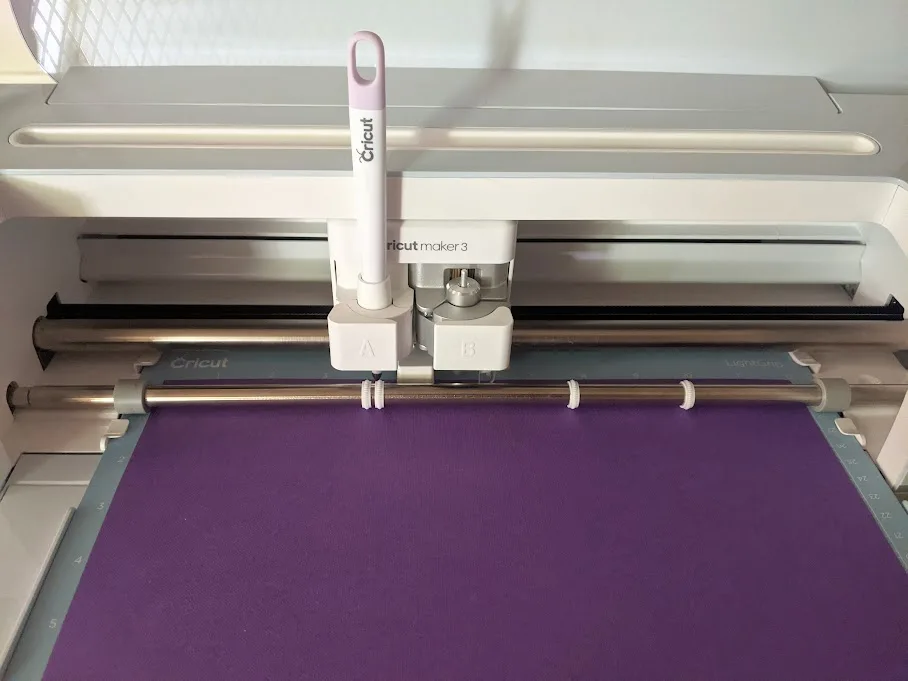 Purple cardstock on machine mat loaded in Cricut Maker 3 and scoring stylus in clamp A.
