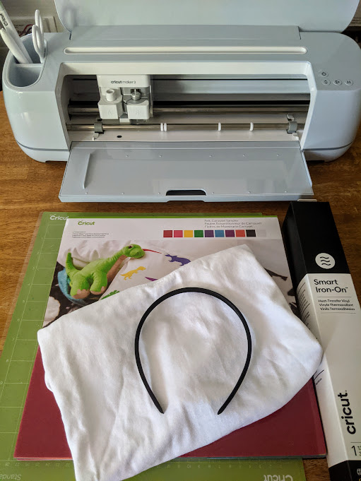 Overhead view of blue Cricut Maker 3, green machine mat, set of Cricut felt, white folded shirt, black headband, and roll of smart black iron on vinyl in box all on the table.