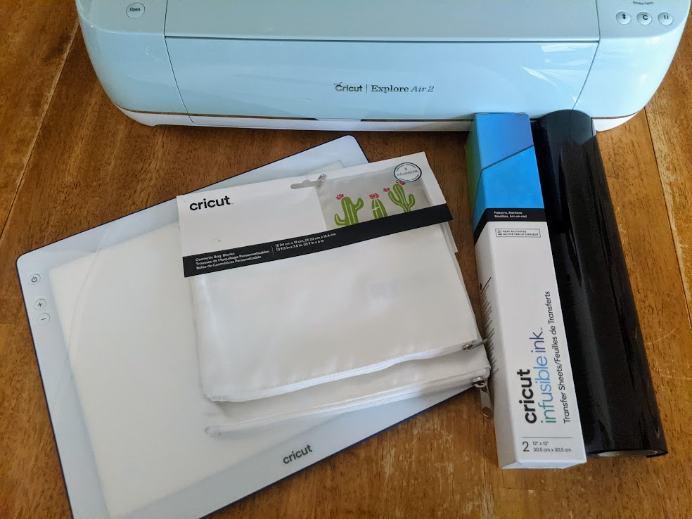 BrightPad Go, Cricut Cosmetic Bag Blanks, Rainbow Infusible Ink Transfer Sheet Box, and roll of Black iron-on vinyl on table in front of mint Cricut Explore Air 2.