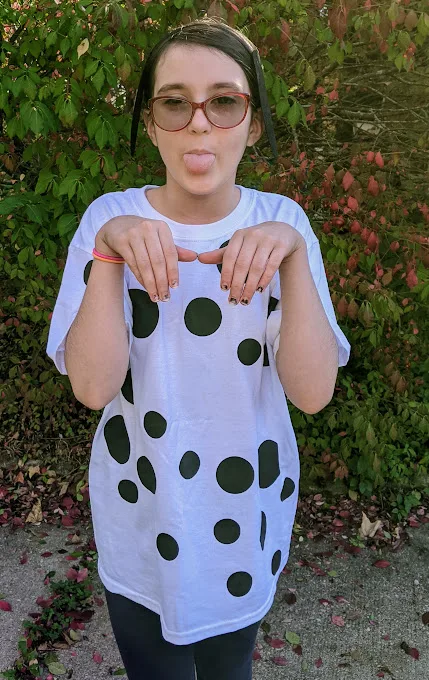 White tween girl wearing finished DIY Dalmatian costume and posing like a dog with tongue sticking out and arms raised with hands curled down like paws.