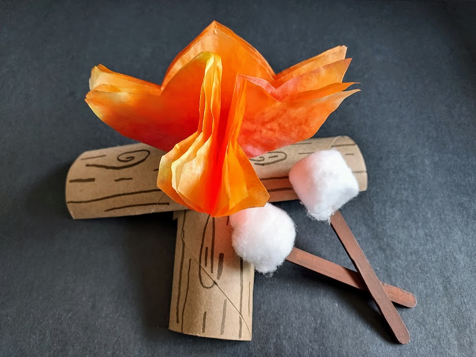 Campfire craft made with orange coffee filter flames and tp tube logs with cotton balls on brown craft sticks to make marshmallows leaning against the campfire.