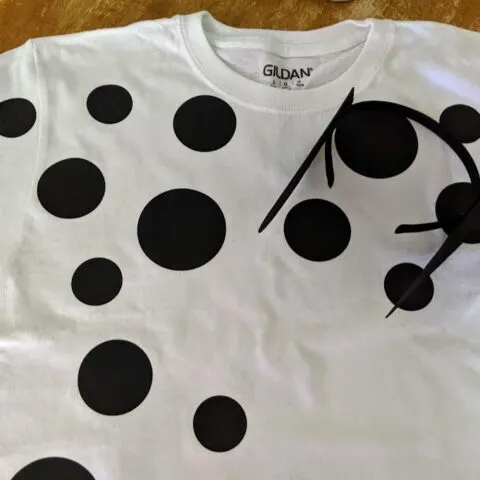 Completed Dalmatian costume: white tshirt with black spots of different sizes ironed on laying out on table. Headband with black felt ears glued on is laying on top of shirt.