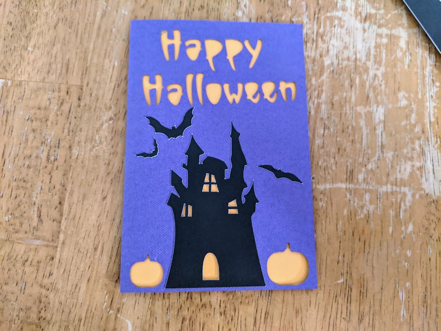 Overhead view of purple Halloween card with Happy Halloween cutout showing orange insert. Pumpkins are cut out to be orange too. Black haunted house silhouette and bats are applied to front of card.