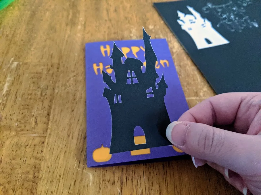 White woman's hand holding haunted house silhouette cut from black sticker cardstock over Halloween card project. 