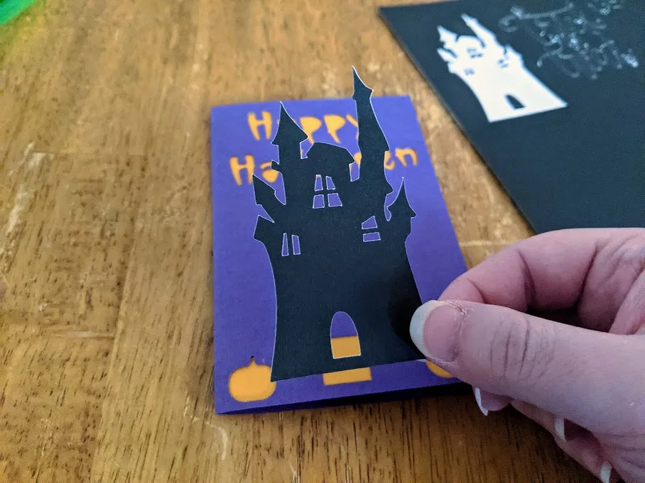 White woman's hand holding haunted house silhouette cut from black sticker cardstock over Halloween card project.
